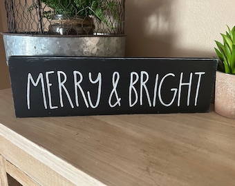 Puede incluir: Letrero de madera negro con las palabras "MERRY & BRIGHT" en letras blancas. El letrero rectangular está sobre una superficie de madera. A la derecha hay una pequeña planta en maceta.