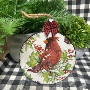 May include: A round, flat ornament featuring a red cardinal bird perched among green holly leaves and red berries. The design includes a dusting of white snow. A small red and black buffalo plaid bow is attached to the top with a jute twine loop.