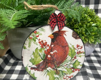 May include: A round, flat ornament featuring a red cardinal bird perched among green holly leaves and red berries. The design includes a dusting of white snow. A small red and black buffalo plaid bow is attached to the top with a jute twine loop.