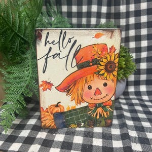 May include: A rectangular wooden sign featuring a scarecrow illustration with a sunflower on its orange hat, a green plaid shirt, and a pumpkin. The text "hello fall" is written in black script. The sign has a distressed finish with autumn leaves.
