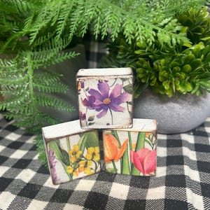 May include: Three small, white wooden boxes with floral designs. One box features a purple flower, another has yellow flowers, and the third displays orange and pink tulips. The boxes are arranged on a black and white checkered surface.
