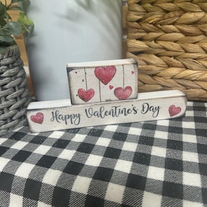 May include: Two small, white wooden blocks with a Valentine's Day theme. The top block features red hearts and the bottom block says "Happy Valentine's Day". The blocks are on a black and white checkered surface.