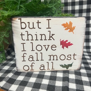 May include: A rectangular wooden sign with a distressed white finish and dark brown edges. The text reads, but I think I love fall most of all, in dark brown lettering. Three painted leaves in orange, red, and green are displayed on the right side.