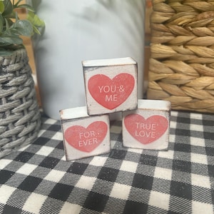 May include: Three small, white wooden blocks with red heart designs. Each block has a different phrase: "YOU & ME", "FOR EVER", and "TRUE LOVE". The blocks are arranged on a black and white checkered surface.