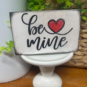 May include: A small, rectangular wooden sign with the words "be mine" in black script and a red glitter heart. The sign has a distressed white background and sits on a white pedestal. The edges of the sign are dark.