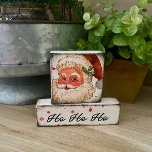 May include: Two small, rectangular wooden blocks. The top block features a vintage image of Santa Claus in a red hat. The bottom block has the phrase "Ho Ho Ho" in black script with red accents. Christmas decor.
