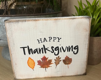 May include: A rectangular, distressed wooden sign with the words "HAPPY Thanksgiving" in black lettering. The sign features four painted fall leaves in orange, red, and brown. The sign is a decorative item for the Thanksgiving holiday.