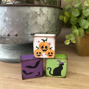 Puede incluir: Tres peque&ntilde;os bloques de madera cuadrados con dise&ntilde;os tem&aacute;ticos de Halloween. Un bloque presenta calabazas naranjas, otro tiene murci&eacute;lagos negros y el tercero muestra un gato negro con un sombrero de bruja. Los bloques est&aacute;n pintados con un acabado envejecido.