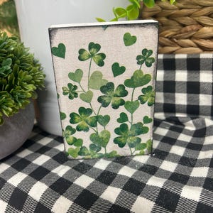 May include: A rectangular, off-white decorative block adorned with green shamrocks and heart-shaped leaves. The block has a slightly distressed look, with a black and white checkered pattern in the background.