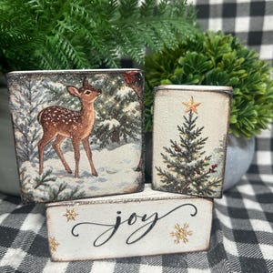 May include: Three rectangular wooden blocks with distressed edges. One block features a brown fawn in a snowy forest, another shows a decorated Christmas tree with a gold star, and the third displays the word joy in black script with gold snowflakes.