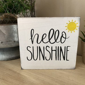 May include: A white wooden block sign with the words "hello SUNSHINE" in black script and a yellow sun graphic. The sign is a decorative item, perfect for adding a touch of cheer to any space.