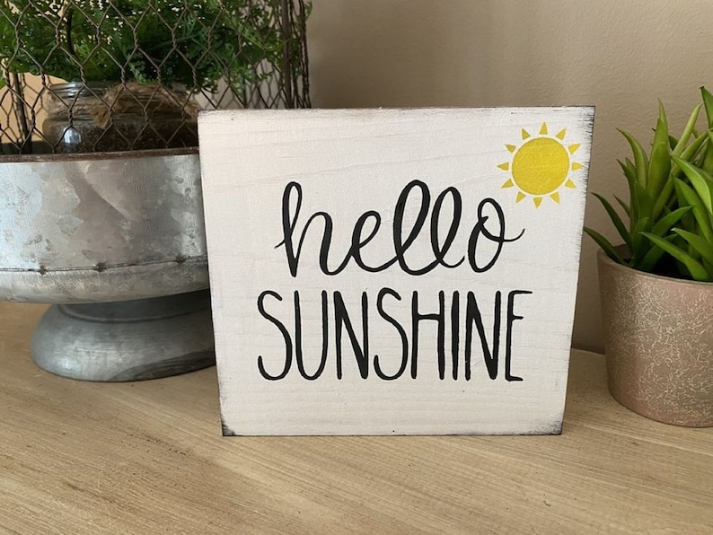 May include: A white wooden block sign with the words "hello SUNSHINE" in black script and a yellow sun graphic. The sign is a decorative item, perfect for adding a touch of cheer to any space.