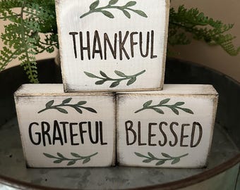 May include: Three white wooden blocks with black lettering that reads "THANKFUL", "GRATEFUL", and "BLESSED". Each block has a green leaf design. The blocks are arranged in a decorative galvanized metal container.