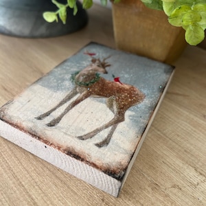 May include: A rectangular wooden block with a vintage illustration of a deer in a snowy scene. The deer has a wreath and red details. The block's edges are lightly burnt, giving a rustic look. A small sprig of greenery is visible in the background.