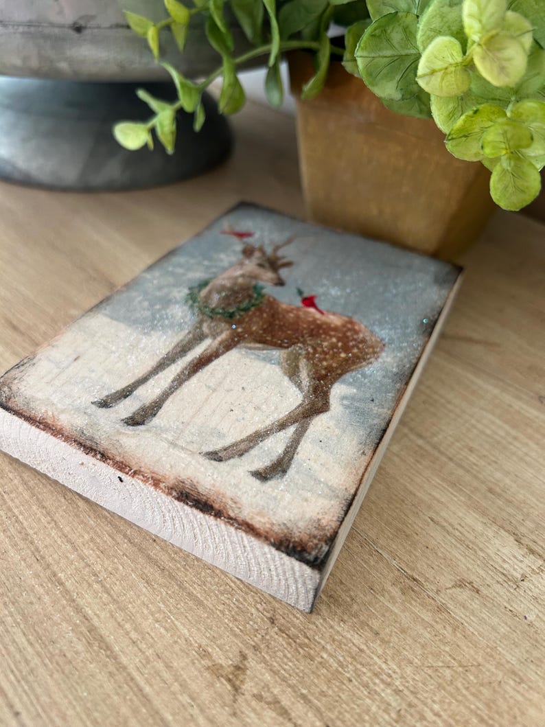 May include: A rectangular wooden block with a vintage illustration of a deer in a snowy scene. The deer has a wreath and red details. The block's edges are lightly burnt, giving a rustic look. A small sprig of greenery is visible in the background.
