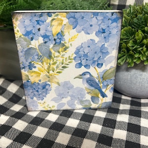 May include: A square planter with a floral design featuring blue hydrangeas and a blue bird. The planter has a white background with blue and yellow accents. The edges of the planter are slightly distressed.