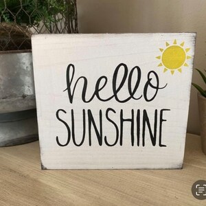 May include: A white wooden block sign with the words "hello SUNSHINE" in black script. A yellow sun graphic is in the upper right corner. The sign is a decorative item.