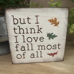 May include: A rustic wooden block sign with the text "but I think I love fall most of all" in dark brown lettering. The sign features three leaf illustrations in orange, green, and red.