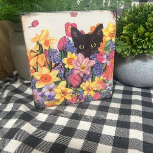 May include: A wooden block featuring a black cat peeking through a vibrant floral arrangement of daffodils, tulips, and other colorful flowers. The artwork has a distressed, vintage look, with a black and white checkered background.