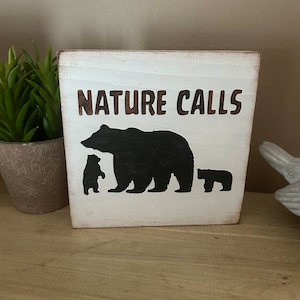 May include: A white wooden block sign with the words "NATURE CALLS" in brown lettering. A black silhouette of a bear and two cubs is below the text. The sign has a rustic, distressed look.