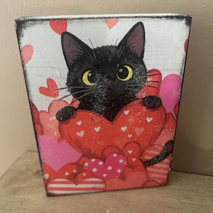 May include: A decorative wooden block featuring a black cat with yellow eyes, holding a large red heart. The block is adorned with various pink and red hearts, creating a Valentine's Day theme. The edges have a distressed, vintage look.