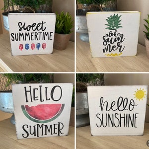 May include: Four white wooden blocks with summer designs. One block says "Sweet Summertime" with popsicle graphics. Another has a pineapple and "Aloha Summer" text. A third says "Hello Summer" with a watermelon. The last says "Hello Sunshine" with a sun.