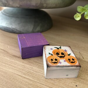 Puede incluir: Dos peque&ntilde;os bloques de madera. Uno est&aacute; pintado de color p&uacute;rpura liso. El otro es blanco con un dise&ntilde;o pintado a mano de tres calabazas naranjas con caras negras y estrellas p&uacute;rpuras, ideal para la decoraci&oacute;n de Halloween.