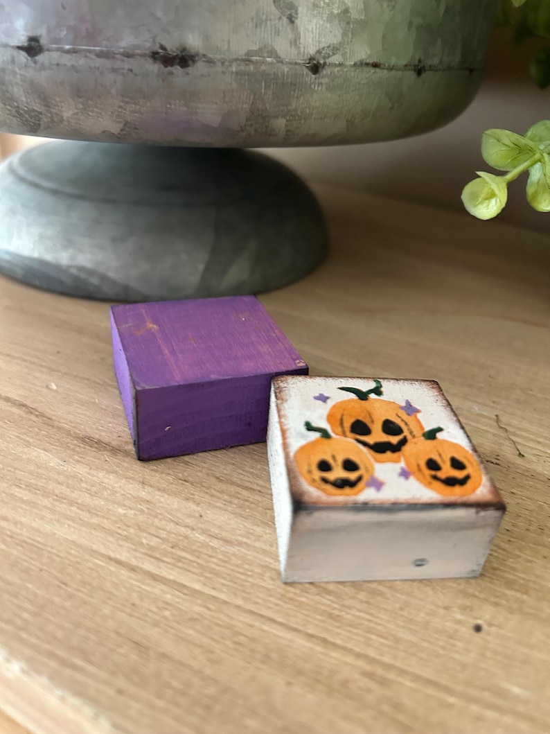 Puede incluir: Dos peque&ntilde;os bloques de madera. Uno est&aacute; pintado de color p&uacute;rpura liso. El otro es blanco con un dise&ntilde;o pintado a mano de tres calabazas naranjas con caras negras y estrellas p&uacute;rpuras, ideal para la decoraci&oacute;n de Halloween.