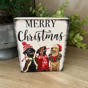 May include: A wooden block with the words "MERRY Christmas" in black script. The block features three dogs wearing Christmas attire: a Santa hat, scarf, and a wreath. The dogs are a black lab, a brown lab, and a golden lab.