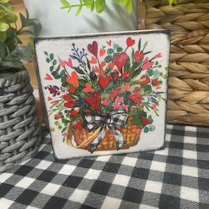 May include: A square wooden block with a floral design featuring red hearts and flowers in a basket with a black and white checkered bow. The block has a distressed, vintage look.