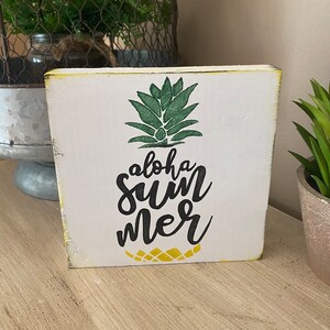 May include: A white wooden block featuring a green pineapple design and the words "aloha summer" in black script. The block has yellowed edges, giving it a rustic appearance. Ideal for a summer or tropical theme.