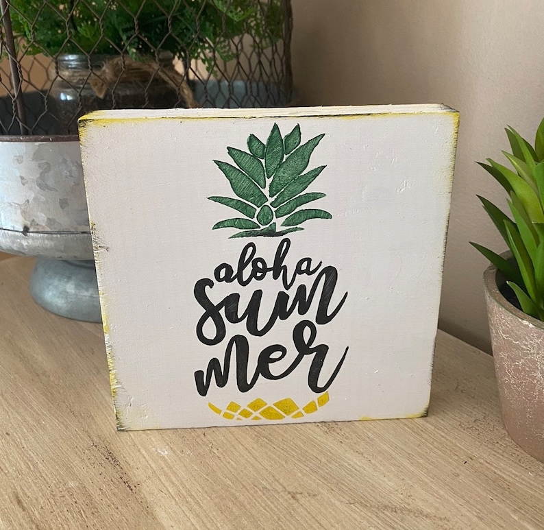 May include: A white wooden block featuring a green pineapple design and the words "aloha summer" in black script. The block has yellowed edges, giving it a rustic appearance. Ideal for a summer or tropical theme.