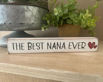 May include: A rectangular, white wooden sign with the text "THE BEST NANA EVER" in black lettering. Two red hearts are painted on the right side. The sign is on a wooden surface.