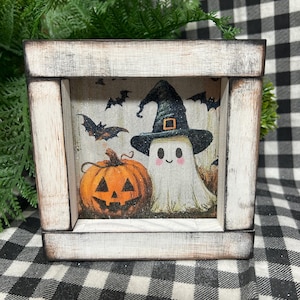 May include: A square wooden sign with a distressed white finish featuring a printed illustration of a smiling ghost wearing a black witch hat next to a carved orange jack-o'-lantern, surrounded by small black bats on a light background.