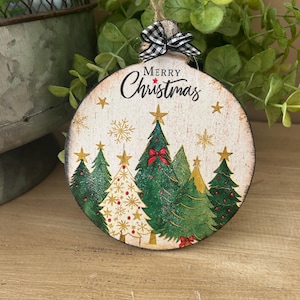 May include: Round wooden Christmas ornament with a rustic design. The ornament features a painted scene of Christmas trees, stars, and the words "Merry Christmas." It is adorned with a black and white checkered bow and a twine loop for hanging.