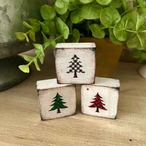 May include: Three small, square, wooden blocks with a distressed white finish. Each block features a different colored Christmas tree design: green, red, and black and white checkered. The blocks are arranged on a wooden surface.