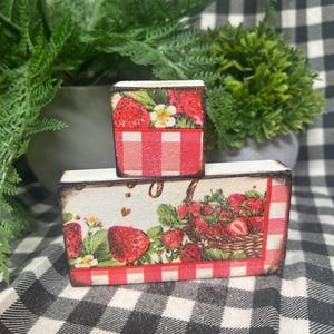 May include: Two rectangular wooden blocks stacked vertically, featuring a decorative print of red strawberries, green leaves, white flowers, and a brown woven basket, accented by a red and white checkered pattern along the bottom edges.