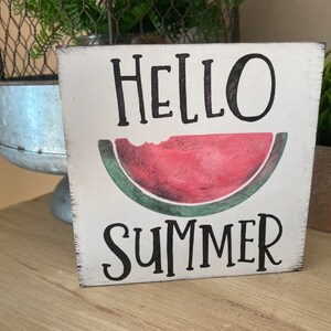 May include: A square, white wooden sign with the words "HELLO SUMMER" in black script. A watercolor-style watermelon slice is in the center. The sign has a distressed, rustic look, ideal for summer decor.