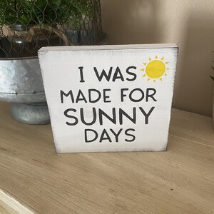 May include: A white wooden block with the text "I WAS MADE FOR SUNNY DAYS" in black and a yellow sun illustration. The block has a distressed finish, suggesting a decorative home accent, possibly for a shelf.