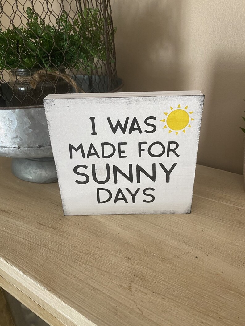 May include: A white wooden block with the text "I WAS MADE FOR SUNNY DAYS" in black and a yellow sun illustration. The block has a distressed finish, suggesting a decorative home accent, possibly for a shelf.