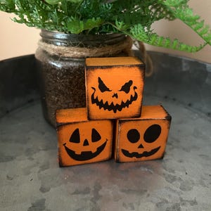 May include: Three orange wooden blocks with black jack-o'-lantern faces. The blocks are stacked on a metal tray, with a glass jar of greenery in the background. The blocks are a Halloween decoration.