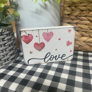 May include: A small, rectangular wooden block with a distressed white finish. It features hand-painted pink and red hearts, gold accents, and the word "love" in cursive. The block is placed on a black and white checkered surface.