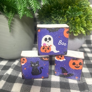 May include: Three square wooden blocks featuring Halloween designs on a purple background. One block shows a white ghost holding an orange pumpkin with the text Boo. Another block displays a black cat and pumpkins. The third block features a witch hat.
