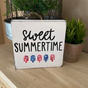 May include: A white wooden block sign with the words "Sweet Summertime" in black script. Below the text are five popsicle illustrations in red and blue. The sign has a distressed, rustic appearance.