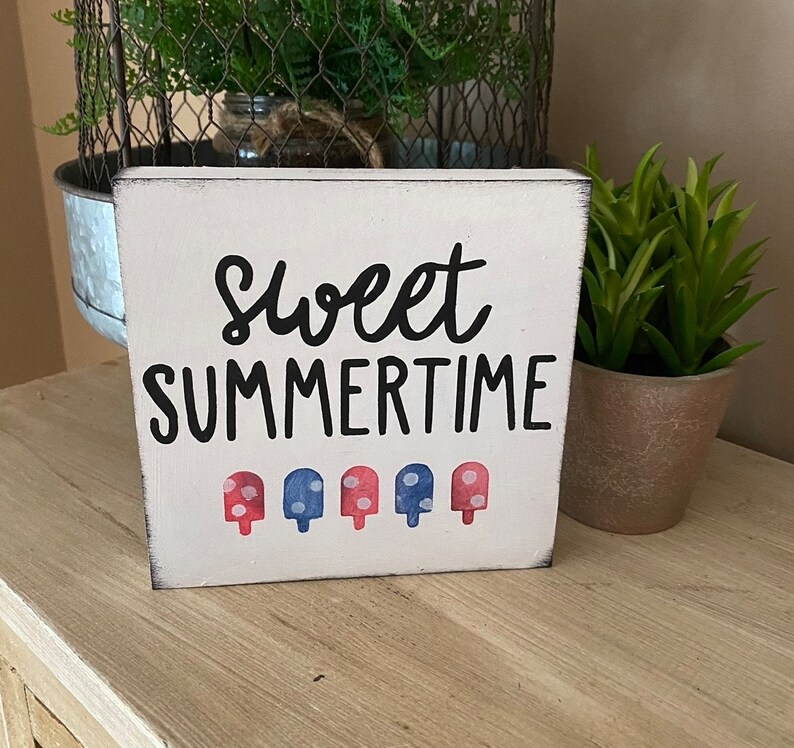 May include: A white wooden block sign with the words "Sweet Summertime" in black script. Below the text are five popsicle illustrations in red and blue. The sign has a distressed, rustic appearance.