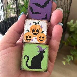Puede incluir: Tres peque&ntilde;os bloques de madera cuadrados apilados, cada uno con un dise&ntilde;o tem&aacute;tico de Halloween. El bloque superior es morado con murci&eacute;lagos negros, el bloque central tiene calabazas naranjas y el bloque inferior es verde con un gato negro con sombrero de bruja.