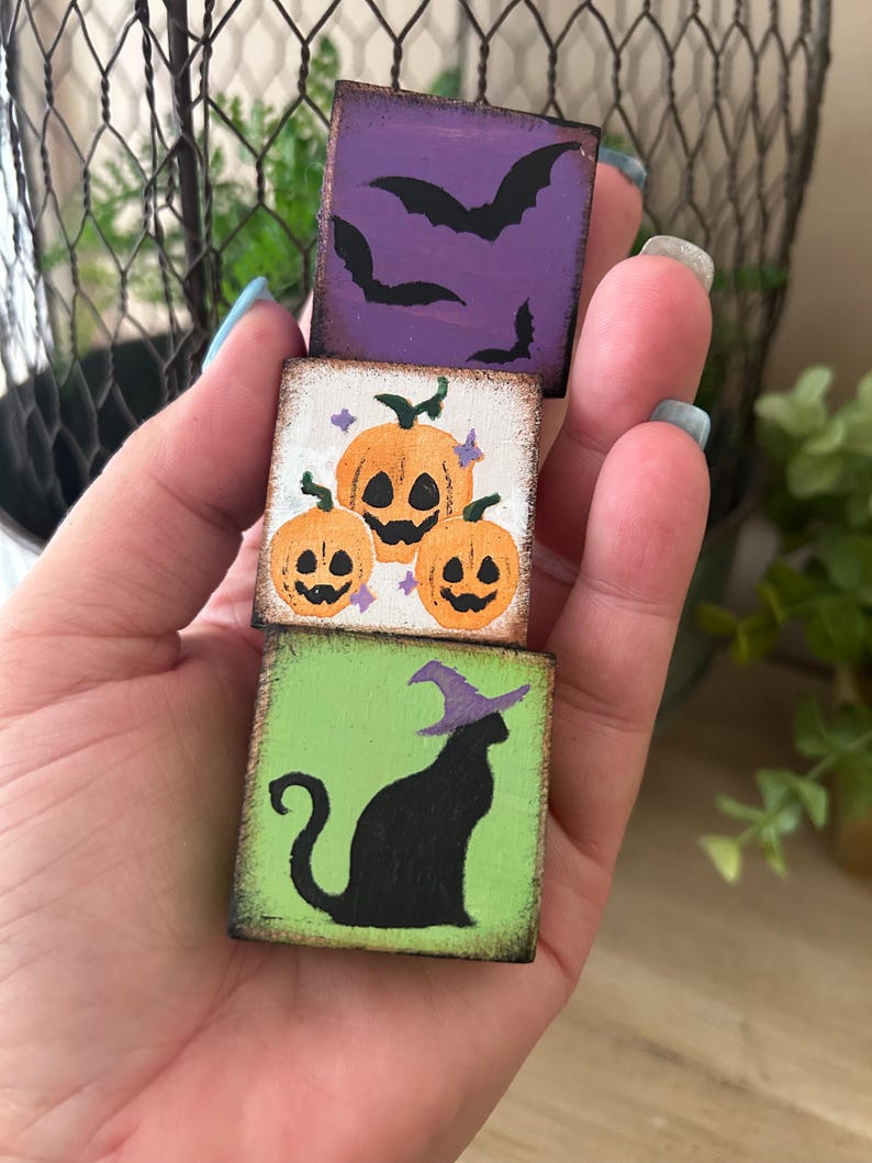 Puede incluir: Tres peque&ntilde;os bloques de madera cuadrados apilados, cada uno con un dise&ntilde;o tem&aacute;tico de Halloween. El bloque superior es morado con murci&eacute;lagos negros, el bloque central tiene calabazas naranjas y el bloque inferior es verde con un gato negro con sombrero de bruja.