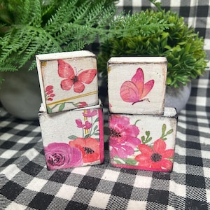 May include: Four small, square wooden blocks with floral and butterfly designs. The blocks feature pink and red flowers and butterflies on a white background. The blocks are arranged on a black and white checkered surface.