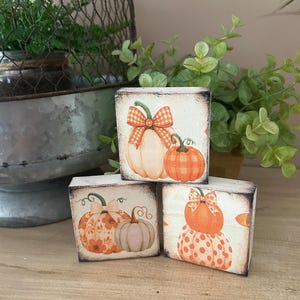 May include: Three wooden blocks with pumpkin designs. Each block features a different pumpkin illustration in shades of orange, cream, and brown. The pumpkins have decorative bows and patterns. The blocks are arranged on a wooden surface.