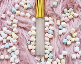 May include: A clear lip gloss tube with a shiny gold cap, filled with a shimmering, translucent liquid. The tube is surrounded by a scattering of small, pastel-coloured marshmallows in shades of pink, yellow, blue, and white.